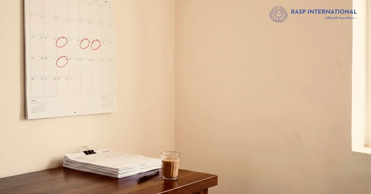 Wall calendar with circled dates above a desk with a stack of papers and a glass of tea